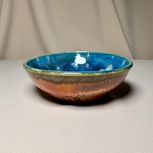 May include: A round ceramic bowl featuring a vibrant blue interior glaze that transitions into a deep green band at the rim. The exterior displays a warm, textured orange and brown gradient finish with visible glaze drips and a smooth, curved shape.