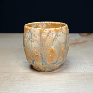 May include: A ceramic cup with a rounded shape and a glossy glaze featuring a blend of warm amber, golden yellow, and soft blue tones. The surface has a textured, mottled appearance with subtle crystalline patterns throughout the exterior.