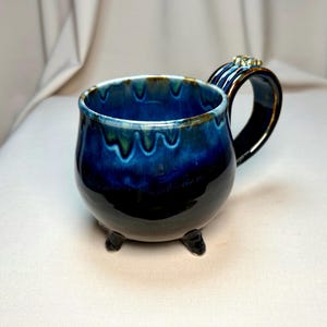 Peut inclure: Une tasse en céramique avec un corps arrondi et trois petits pieds. L'extérieur présente une glaçure noire profonde qui se transforme en un motif de gouttes bleu et vert vif près du bord. Elle possède une anse incurvée avec un petit ornement.