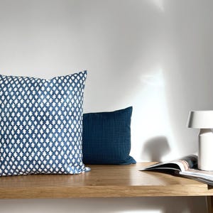 Könnte beinhalten: Zwei dekorative Kissen auf einem Holzregal. Ein Kissen ist marineblau mit einem weißen geometrischen Muster, das andere ist einfarbig marineblau. Eine weiße Lampe und eine offene Zeitschrift befinden sich ebenfalls auf dem Regal.