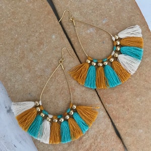 May include: A pair of gold hoop earrings with a teardrop shape. The earrings are adorned with a fringe of white, mustard yellow, and teal tassels.