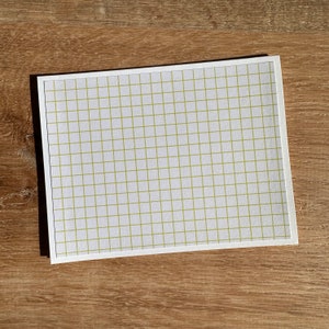 May include: A white rectangular card with a grid pattern in a light green color. The card is on a wooden surface, with the shadow of the card visible. The grid pattern is evenly spaced.