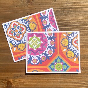 May include: Two greeting cards with a colorful, patterned design. The cards feature a repeating geometric pattern in shades of pink, orange, blue, and yellow, with a white border. The cards are displayed on a wooden surface.