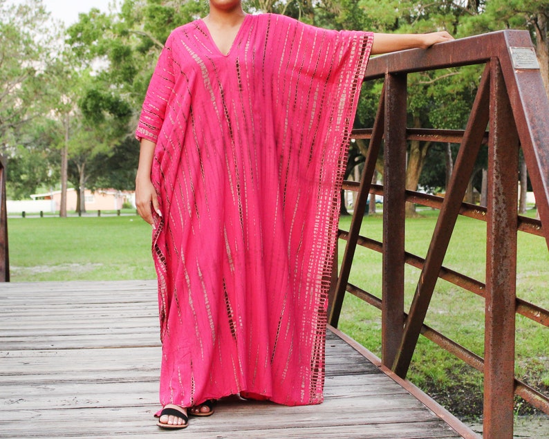K&ouml;nnte beinhalten: Ein pinkfarbenes und braunes Batik-Kaftan mit V-Ausschnitt. Der Kaftan ist lang und flie&szlig;end und wird von einer Person getragen, die auf einer Holzbr&uuml;cke steht.