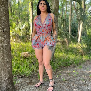 May include: A woman wearing a blue and red floral print romper with a V-neckline and a tie at the waist. She is standing in a wooded area.