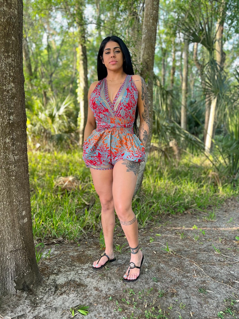 May include: A woman wearing a blue and red floral print romper with a V-neckline and a tie at the waist. She is standing in a wooded area.