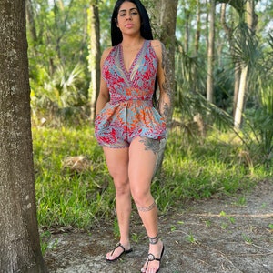 May include: A woman wearing a blue and red floral print romper with a V-neckline and a tie at the waist. She is standing in a wooded area.
