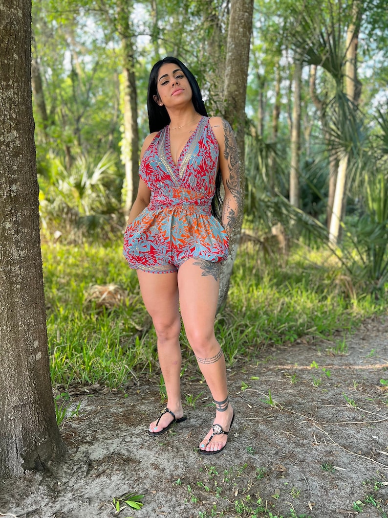 May include: A woman wearing a red and blue floral print jumpsuit with a V-neck and a tie at the waist. She is standing in a wooded area with trees in the background.
