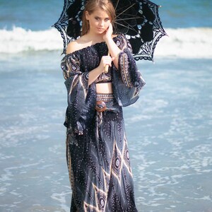 May include: A woman wearing a black and gold patterned dress and a black lace parasol stands in the shallow water of the ocean.