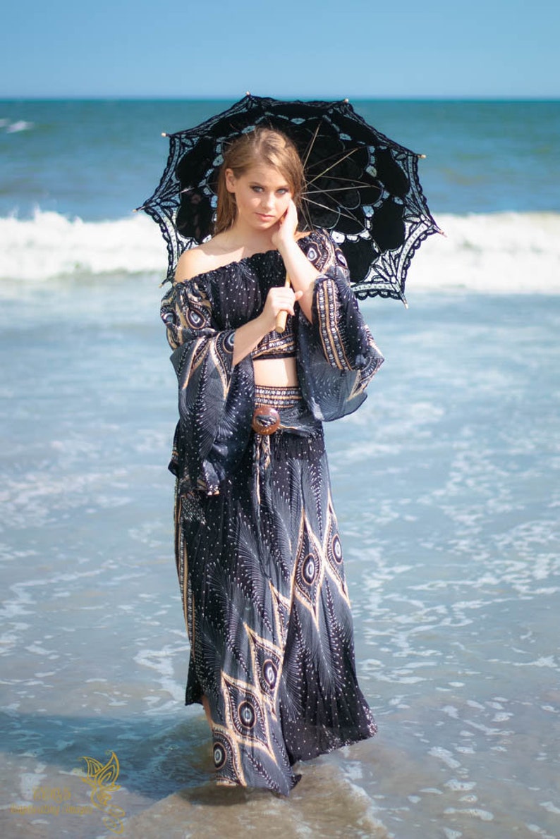 May include: A woman wearing a black and gold patterned dress and a black lace parasol stands in the shallow water of the ocean.