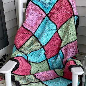 May include: A crocheted throw blanket with a diamond pattern in shades of pink, blue, green, and black. The blanket is draped over a white rocking chair on a porch.