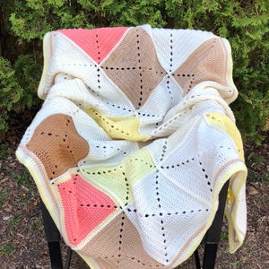 Puede incluir: Una manta de ganchillo con un diseño de patchwork en tonos de blanco, melocotón, amarillo y marrón. La manta está doblada y drapeada sobre una silla de metal negro.