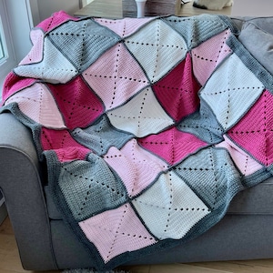 May include: A crocheted blanket with a patchwork design in shades of pink, grey, and white. The blanket is made of squares with a diamond pattern.