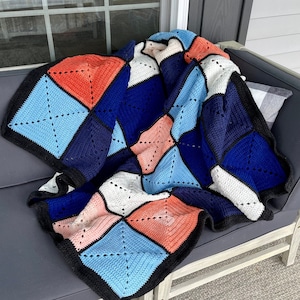 Puede incluir: Una manta de ganchillo con un patrón geométrico en tonos de azul, rosa y blanco. La manta está doblada y colocada sobre un sofá gris.