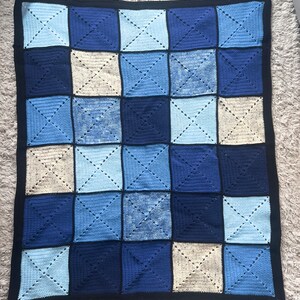 以下が含まれることがあります： 青とベージュのチェック柄の、手編みのベビーブランケット。柔らかな糸で作られており、黒い縁取りが施されています。