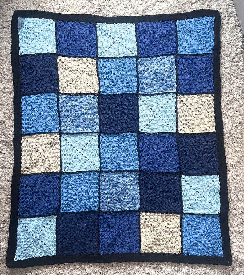 以下が含まれることがあります： 青とベージュのチェック柄の、手編みのベビーブランケット。柔らかな糸で作られており、黒い縁取りが施されています。