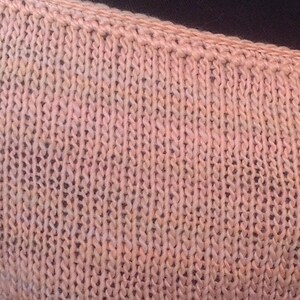 May include: Close-up of a knitted fabric in a pale pink color with a subtle variegated pattern. The fabric has a smooth texture and a slightly textured surface.