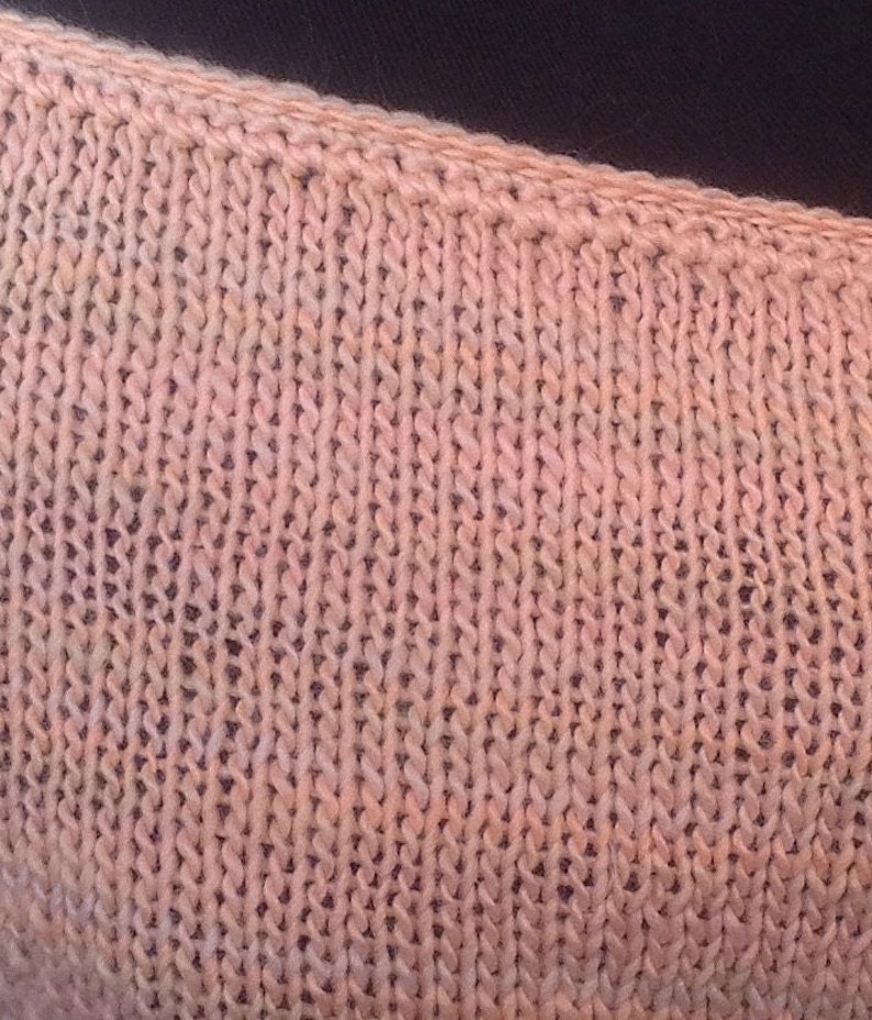 May include: Close-up of a knitted fabric in a pale pink color with a subtle variegated pattern. The fabric has a smooth texture and a slightly textured surface.