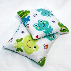 May include: Two white fabric pillow covers with a green frog pattern. The pillows are stacked on top of each other. The top pillow has a blue frog with a star and a bee on it. The bottom pillow has a green frog with a star and a bee on it.
