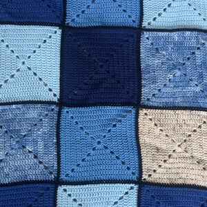 以下が含まれることがあります： 青とベージュの色合いの正方形でできた、かぎ針編みの毛布。正方形は濃い青色の糸でつなぎ合わされています。