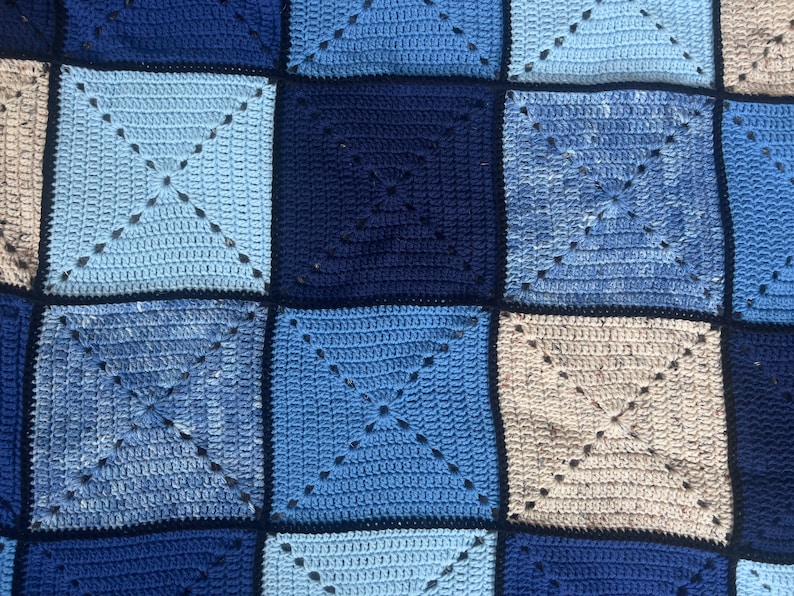 以下が含まれることがあります： 青とベージュの色合いの正方形でできた、かぎ針編みの毛布。正方形は濃い青色の糸でつなぎ合わされています。