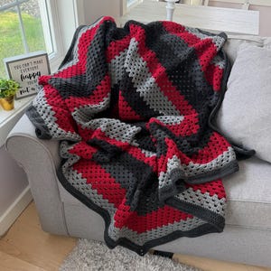 Könnte beinhalten: Eine handgefertigte Häkeldecke mit Zickzackmuster in abwechselnden Streifen aus anthrazitfarbenem, hellrotem und hellgrauem Garn. Die Decke hat ein strukturiertes Design mit offenen Maschen und liegt über einem grauen Sofa.