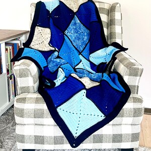 以下が含まれることがあります： グレーと白のチェック柄の肘掛け椅子に、青、白、グレーのダイヤモンド柄のクロシェ編み毛布がかけられています。