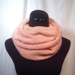 May include: A soft, peach-colored knitted infinity scarf. The scarf is folded and displayed on a black mannequin.