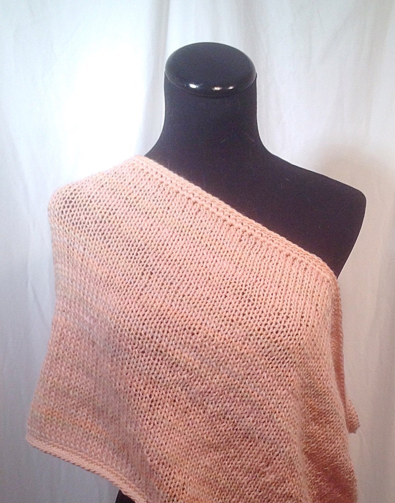 May include: A peach-colored knitted shawl with a one-shoulder design. The shawl is draped over a black mannequin.