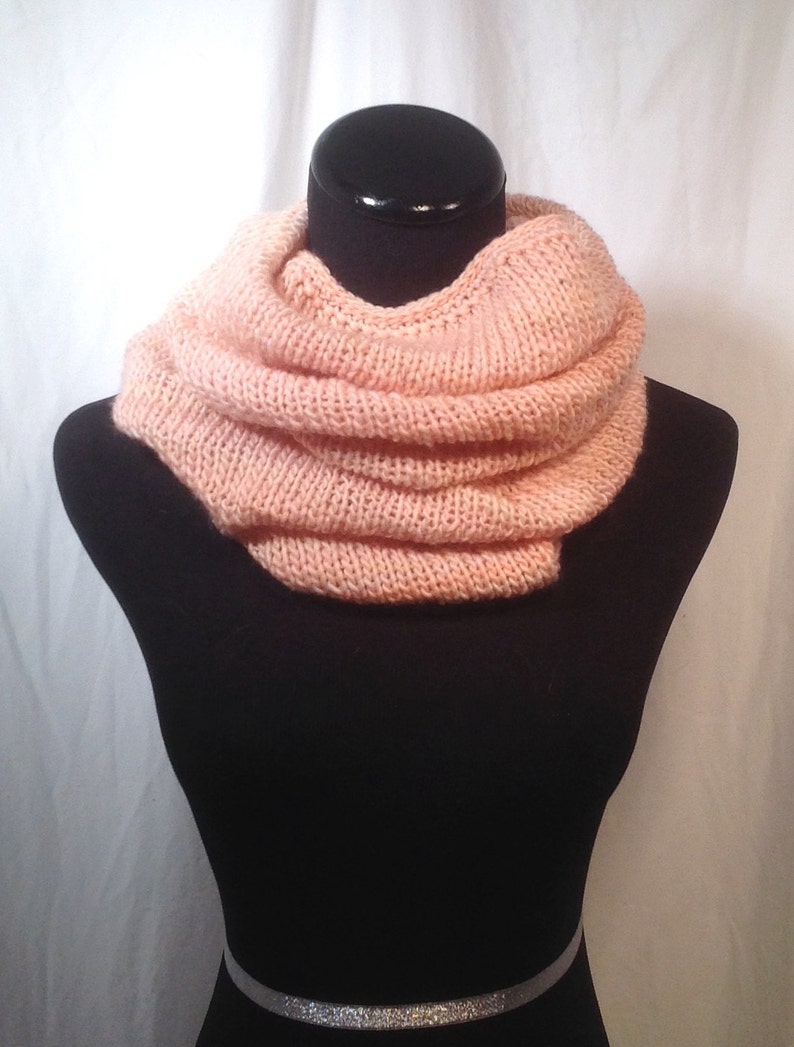 May include: A soft, peach-colored knitted infinity scarf. The scarf is folded and displayed on a black mannequin.