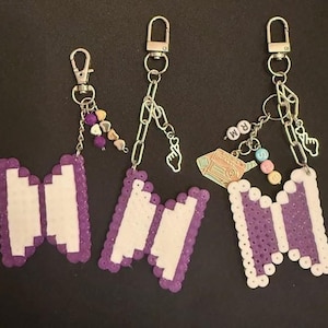 May include: Three keychains with the BTS logo in purple and white, made of small beads. Each keychain has a silver clasp and chain, with decorative beads and charms.