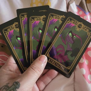 May include: Five tarot cards with black backgrounds and gold trim. The cards feature a green and purple creature with glowing green eyes. The card on top reads "STRENGTH" in gold lettering.