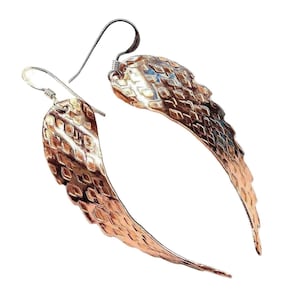 May include: A pair of copper-colored wing-shaped earrings. The earrings have a textured surface with a diamond pattern. The ear wires are silver-colored. The earrings are displayed against a white background.