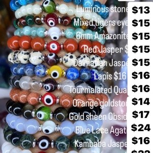 K&ouml;nnte beinhalten: Ein Stapel von zehn Armb&auml;ndern mit verschiedenen farbigen Steinen und silbernen b&ouml;sen Augen-Anh&auml;ngern. Die Armb&auml;nder sind mit der Art des Steins und dem Preis gekennzeichnet. Apatit 17$, Leuchtender Stein 13$, Gemischtes Tigerauge 15$, 6mm Amazonit 15$, Roter Jaspis 15$, Dalmatiner Jaspis 15$, Lapislazuli 16$ 16$, Tourmalinierter Quarz 16$, Orange Goldstone 14$, Goldschimmer Obsidian 17$, Blaue Spitzen-Achat 24$, Kambaba Jaspis 16$, Mondstein 22$, 8mm Amazonit 19$.