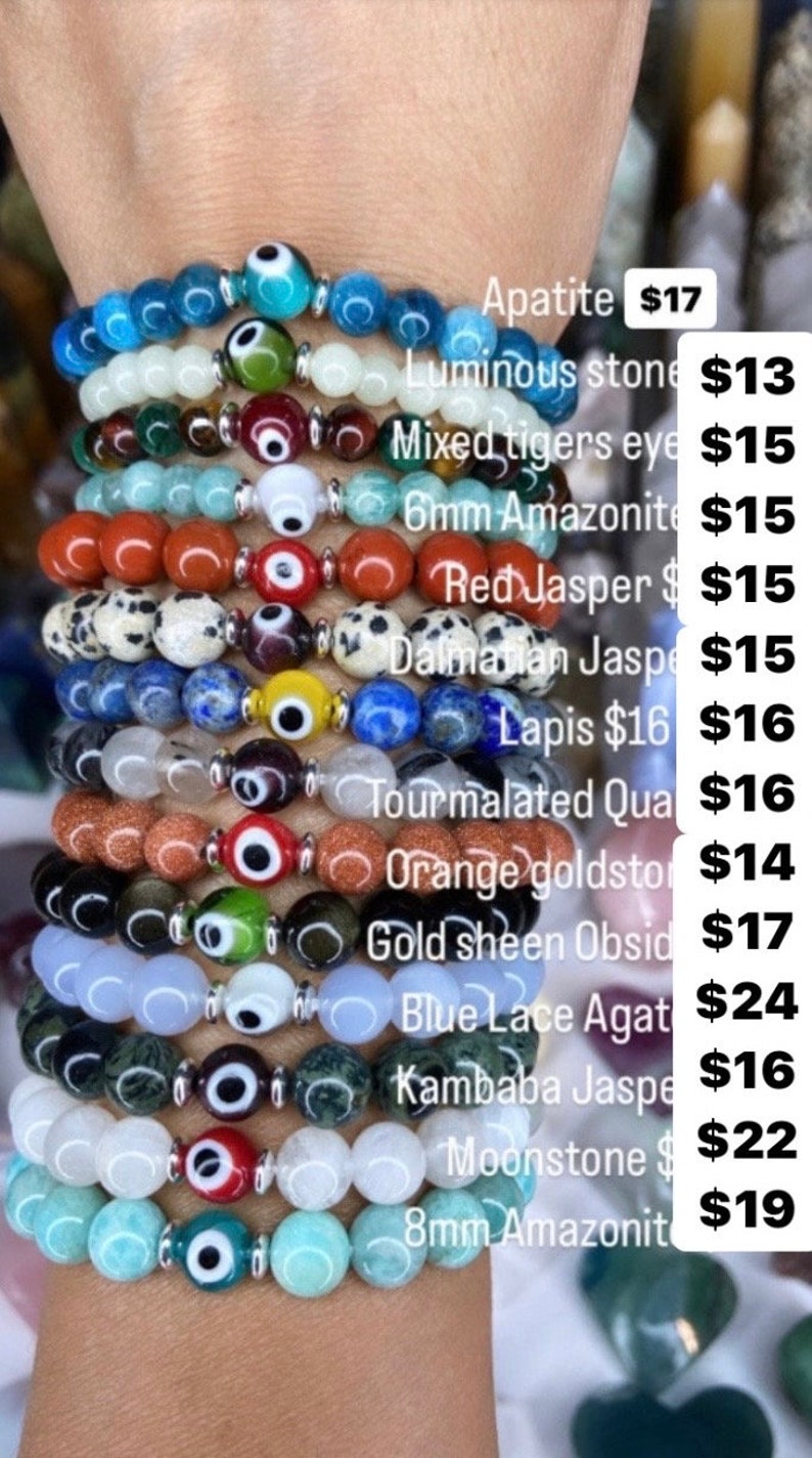 K&ouml;nnte beinhalten: Ein Stapel von zehn Armb&auml;ndern mit verschiedenen farbigen Steinen und silbernen b&ouml;sen Augen-Anh&auml;ngern. Die Armb&auml;nder sind mit der Art des Steins und dem Preis gekennzeichnet. Apatit 17$, Leuchtender Stein 13$, Gemischtes Tigerauge 15$, 6mm Amazonit 15$, Roter Jaspis 15$, Dalmatiner Jaspis 15$, Lapislazuli 16$ 16$, Tourmalinierter Quarz 16$, Orange Goldstone 14$, Goldschimmer Obsidian 17$, Blaue Spitzen-Achat 24$, Kambaba Jaspis 16$, Mondstein 22$, 8mm Amazonit 19$.