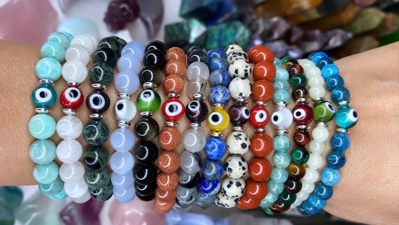 K&ouml;nnte beinhalten: Ein Stapel von 14 Armb&auml;ndern mit verschiedenen Edelsteinperlen und b&ouml;sen Augen-Anh&auml;ngern. Die Armb&auml;nder sind in Blaut&ouml;nen, Gr&uuml;n, Rot, Wei&szlig;, Schwarz und Braun.