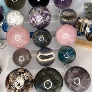 May include: A collection of 15 polished crystal spheres in various colors and textures, including amethyst, rose quartz, labradorite, and agate. The spheres are displayed on a white surface.
