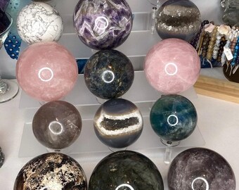 May include: A collection of 15 polished crystal spheres in various colors and textures, including amethyst, rose quartz, labradorite, and agate. The spheres are displayed on a white surface.