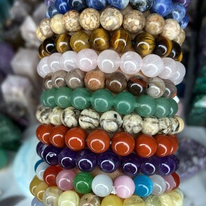 K&ouml;nnte beinhalten: Ein Stapel von 14 bunten Perlenarmb&auml;ndern, jedes aus verschiedenen Steinsorten gefertigt. Die Armb&auml;nder sind &uuml;bereinander auf einem Handgelenk gelegt, beginnend mit einem violetten Amethyst-Armband und endend mit einem braunen und wei&szlig;en gestreiften Armband.