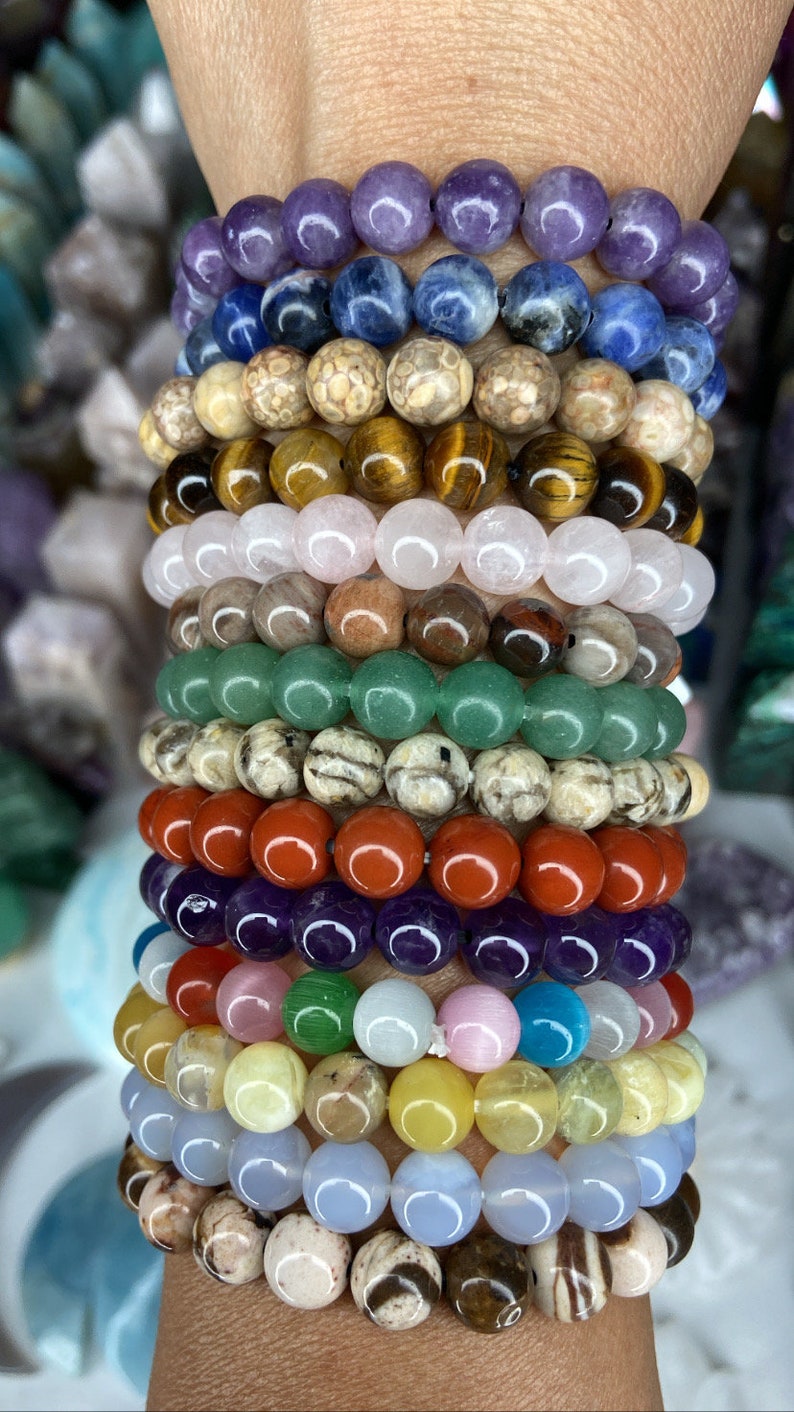 K&ouml;nnte beinhalten: Ein Stapel von 14 bunten Perlenarmb&auml;ndern, jedes aus verschiedenen Steinsorten gefertigt. Die Armb&auml;nder sind &uuml;bereinander auf einem Handgelenk gelegt, beginnend mit einem violetten Amethyst-Armband und endend mit einem braunen und wei&szlig;en gestreiften Armband.