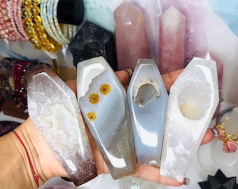 May include: Four polished agate crystals shaped like coffins held in a hand. The stones feature varied textures including translucent bands, crystalline druzy centers, and small circular inclusions in shades of white, gray, and brown.