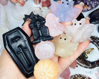 May include: A collection of small carved stone figures held in a palm, including a black coffin, a black tombstone, a light blue bat, a pink ghost, two light-colored bats, and two small round pumpkin-shaped carvings in pink and orange.