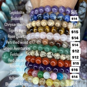 K&ouml;nnte beinhalten: Ein Stapel von 12 Armb&auml;ndern aus verschiedenen Edelsteinen, darunter Amethyst, Rosenquarz, Tigerauge und gr&uuml;ner Aventurin. Jedes Armband ist mit der Art des Steins und dem Preis gekennzeichnet.