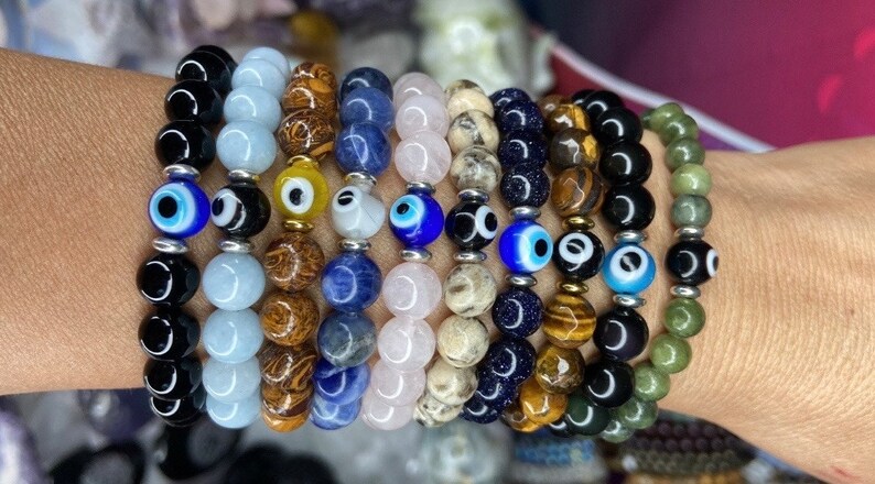 K&ouml;nnte beinhalten: Ein Stapel von acht Armb&auml;ndern mit verschiedenen farbigen Perlen und b&ouml;sen Augen-Anh&auml;ngern. Die Armb&auml;nder sind aus verschiedenen Materialien gefertigt, darunter schwarzer Onyx, Aquamarin, Tigerauge, Lapislazuli, Rosenquarz und gr&uuml;ner Aventurin.