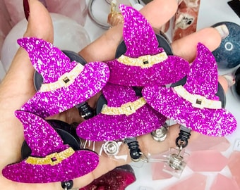 May include: A collection of four identical badge reels, each featuring a bright purple glitter witch hat with a gold glitter band and a small black buckle detail. Each hat is attached to a circular black plastic retractable badge reel mechanism.