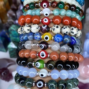 K&ouml;nnte beinhalten: Ein Stapel von 12 Armb&auml;ndern mit unterschiedlich farbigen Perlen und silbernen Gl&uuml;cksbringer-Anh&auml;ngern in Form eines b&ouml;sen Auges. Die Armb&auml;nder sind auf dem Handgelenk einer Person gestapelt.