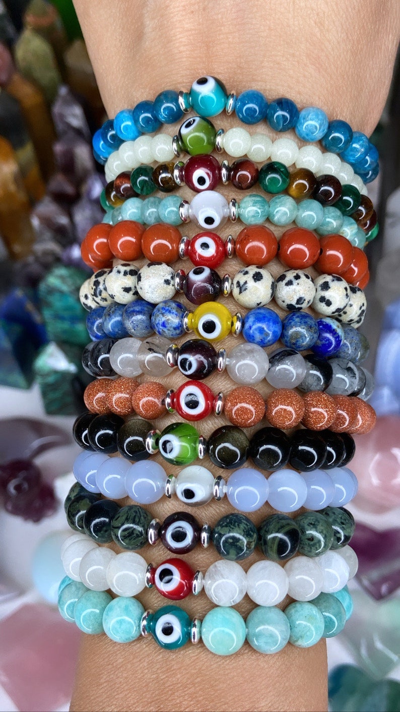 K&ouml;nnte beinhalten: Ein Stapel von 12 Armb&auml;ndern mit unterschiedlich farbigen Perlen und silbernen Gl&uuml;cksbringer-Anh&auml;ngern in Form eines b&ouml;sen Auges. Die Armb&auml;nder sind auf dem Handgelenk einer Person gestapelt.
