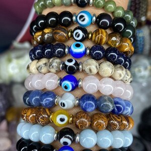 K&ouml;nnte beinhalten: Ein Stapel von zehn Armb&auml;ndern mit blauen b&ouml;sen Augen-Anh&auml;ngern. Die Armb&auml;nder bestehen aus verschiedenen Materialien, darunter schwarzer Onyx, Tigerauge, gr&uuml;ner Jade, blauer Lapislazuli, Rosenquarz und Aquamarin.