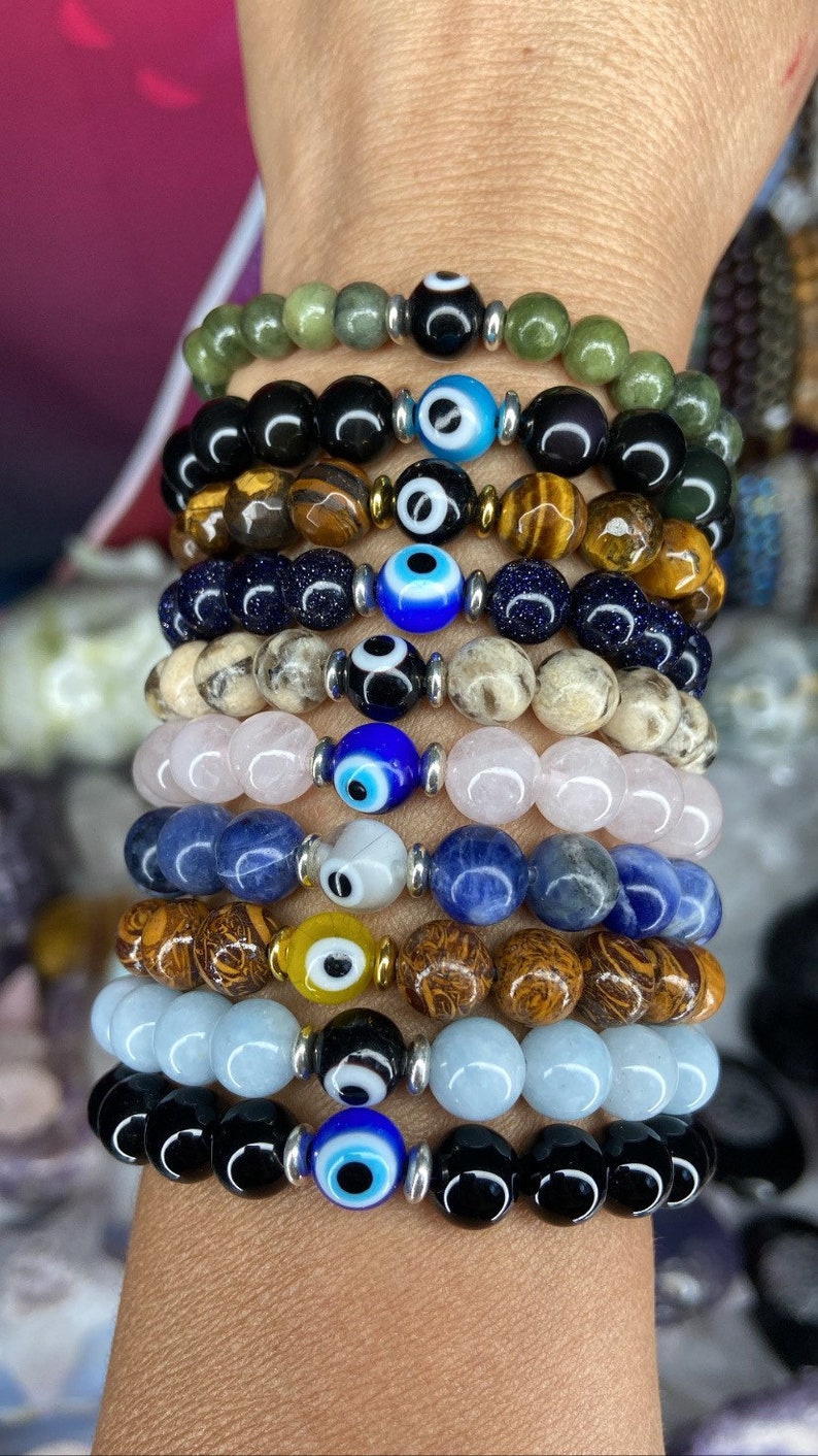 K&ouml;nnte beinhalten: Ein Stapel von zehn Armb&auml;ndern mit blauen b&ouml;sen Augen-Anh&auml;ngern. Die Armb&auml;nder bestehen aus verschiedenen Materialien, darunter schwarzer Onyx, Tigerauge, gr&uuml;ner Jade, blauer Lapislazuli, Rosenquarz und Aquamarin.