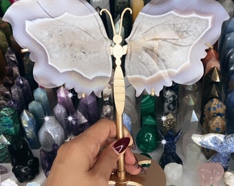May include: A decorative butterfly sculpture with agate wings and a gold-colored metal stand. The wings are a light gray with white and brown patterns. The sculpture is held in front of a collection of crystals and gemstones.
